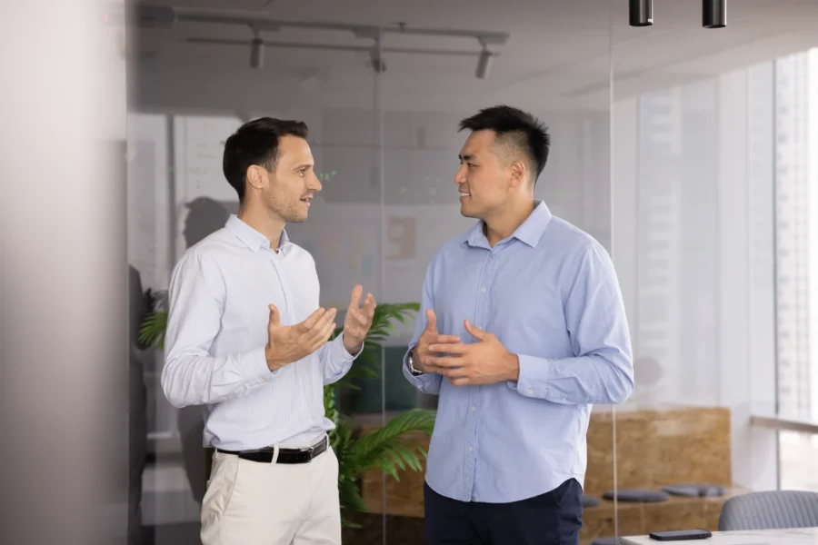 Teammates talking standing in office, smiling, collaborating on creative ideas and strategy, giving feedback on recent joint project completion or work tasks, provide explanation, engaging in teamwork