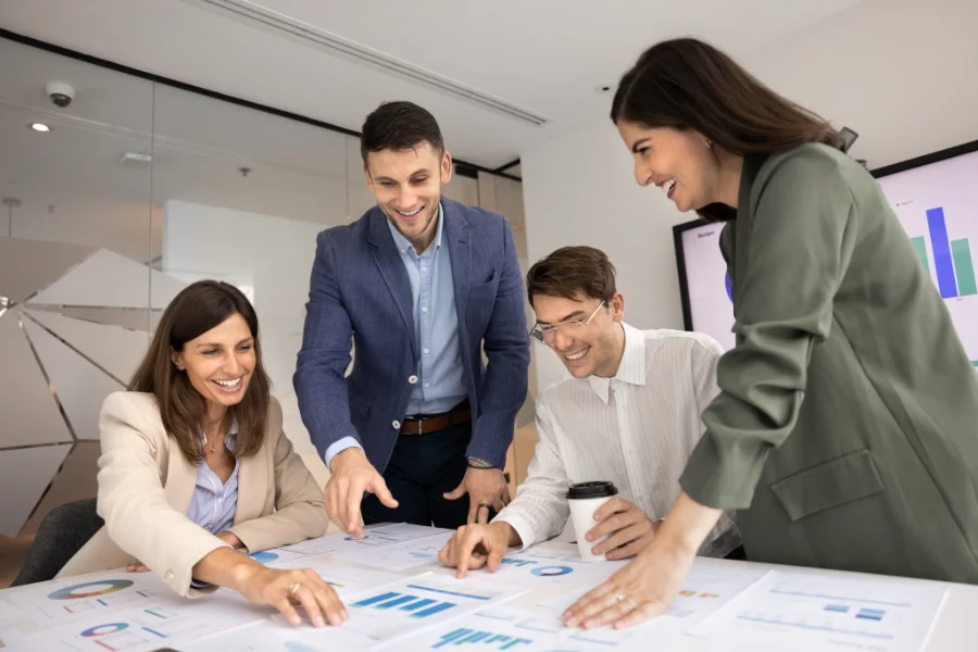 Team of four happy agency coworkers analyzing paper marketing reports on meeting table, pointing at charts, smiling, laughing, discussing ideas for successful startup strategy, sales growth