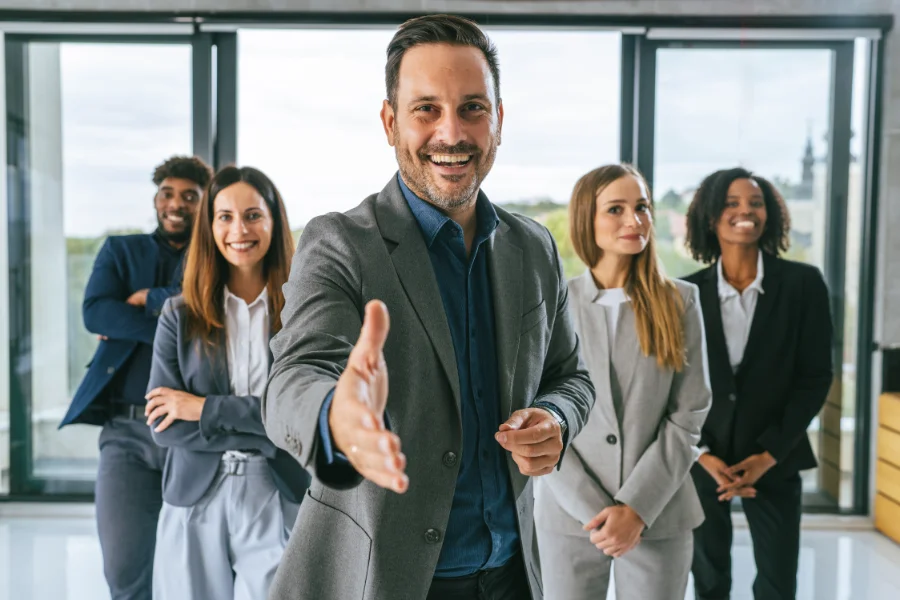 Successful business executive smiling at camera and extending a hand, welcoming or greeting a diverse team of professionals standing confidently in a modern office environment