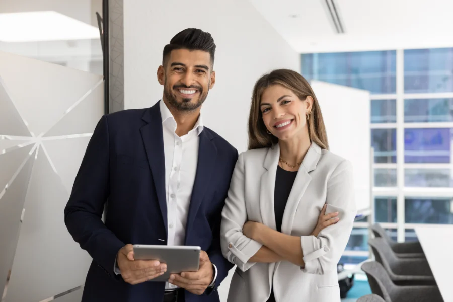 Diverse couple of happy coworkers standing close in office space, posing together, holding tablet pc, looking at camera with toothy smiles, enjoying teamwork, corporate friendship