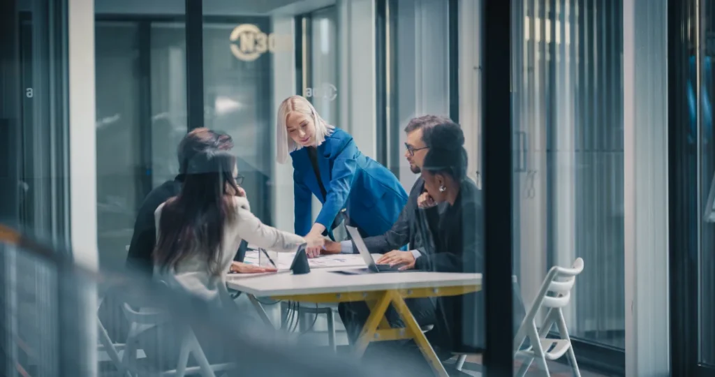 Female Manager Leads a Focused Business Meeting in a Glass Office, Pointing at Documents on the Table While the Team Reviews Charts, Aligns Priorities, Shares Ideas, and Collaborates on Laptops